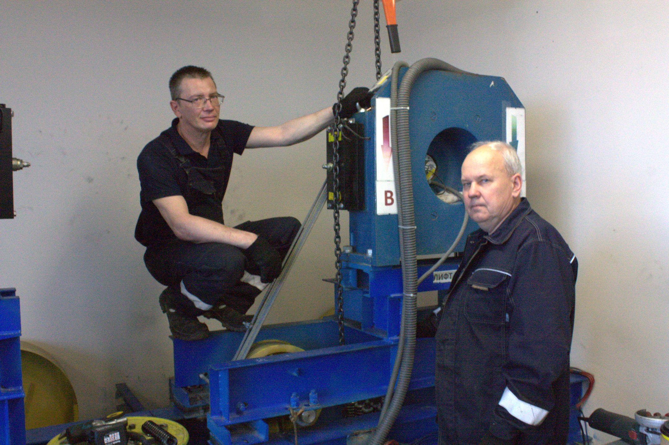 На изображении изображены двое мужчин в мастерской или на производстве, которые, вероятно, занимаются обслуживанием или установкой оборудования. Оба мужчины одеты в тёмную практичную рабочую одежду, в том числе в куртки и перчатки, что говорит о том, что они выполняют технические или механические работы. Мужчина слева стоит на коленях или сидит на корточках рядом с большой синей машиной, положив руку на механизм. Он выглядит сосредоточенным и чувствует себя комфортно рядом с оборудованием. На нём чёрная рубашка и тёмные брюки, он носит очки, что добавляет деталей к его профессиональному образу. У мужчины справа, который, по-видимому, стоит, более серьёзное выражение лица, возможно, указывающее на сосредоточенность или важность выполняемой задачи. Его светлые волосы контрастируют с тёмной рабочей курткой со светоотражающими или усиленными нашивками, указывающими на то, что одежда предназначена для обеспечения безопасности на производстве. Машина, с которой они работают, массивная, окрашена в синий цвет и, по-видимому, является частью более крупной установки, возможно, промышленного двигателя, редуктора или компонента механической системы. Машина включает в себя видимые элементы, такие как цепи, шкив и трубки или шланги, что указывает на сложную механическую или гидравлическую систему. На машине есть стрелки, указывающие направление, и надписи, что говорит о том, что она спроектирована с учётом рекомендаций по эксплуатации, возможно, для облегчения устранения неполадок или технического обслуживания. Сцена ярко освещена, а простой фон указывает на то, что действие происходит в помещении, где основное внимание уделяется функциональности, например, в комнате техобслуживания или аппаратной, а не в цехе или на заводе. Поза и сосредоточенность мужчин говорят о том, что они либо настраивают, ремонтируют или осматривают машину, выполняя задачу, требующую технических навыков и осведомлённости. Инструменты и детали в нижней части кадра, например, чёрное устройство с красной кнопкой, усиливают впечатление от продолжающейся технической работы. В целом, изображение передаёт ощущение профессионализма, рутинности и физической нагрузки, связанных с обслуживанием или установкой промышленного оборудования.