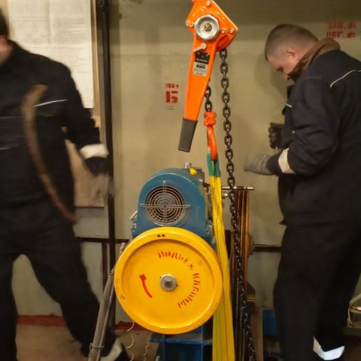 На этом изображении, запечатлены работы по техническому обслуживанию и ремонту лифта в промышленной шахте, в шахте лифта или вспомогательной шахте внутри большого здания. Присутствие человека подразумевает активную работу или осмотр в этом ограниченном пространстве, что может быть опасно и подчёркивает необходимость соблюдения осторожности в таких условиях. В целом, эта фотография даёт представление о работе на производстве, используя перспективу, цвет и структуру, чтобы подчеркнуть высоту и узкость пространства, а также сосредоточенность, необходимую для выполнения задач в этом пространстве. В результате получается изображение, которое передаёт масштаб окружающей среды и интенсивность работы.