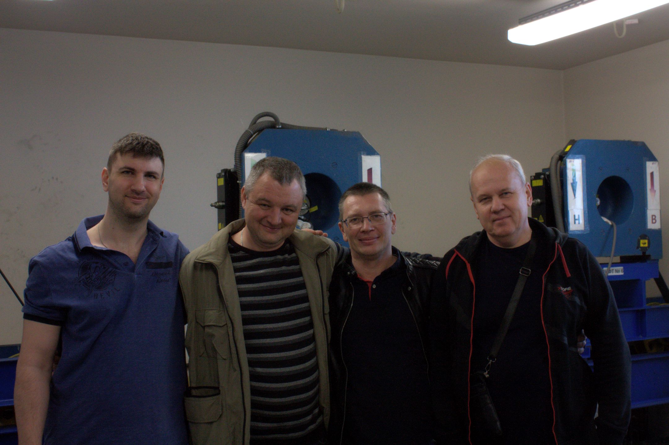 На этом снимке четверо мужчин стоят бок о бок. Кажется, что они позируют в помещении, которое может иметь промышленное или техническое назначение. Фон состоит из машины синего цвета с несколькими панелями, указывающими направление. Мужчины были одеты в повседневную одежду; на одном из них была синяя футболка, в то время как на другом были куртка и брюки разных цветов. Все улыбались и казались знакомыми друг другу. В комнате довольно хорошее освещение, что свидетельствует о приятной атмосфере.