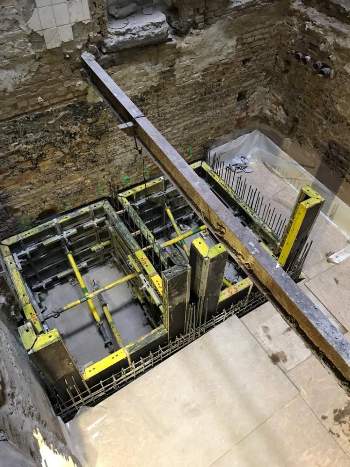 На этом изображении показана строительная площадка внутри помещения из кирпича или бетона. В центре изображения коробчатая конструкция, которая выглядит как устанавливаемая бетонная опалубка. Эта опалубка окружена стальной арматурой, которая заметно выступает наружу. Поверх таких конструкций устанавливаются длинные балки, которые могут использоваться для поддержки или подъема материала. Стены вокруг них кажутся шероховатыми и текстурированными, указывая на то, что эта комната находится в процессе ремонта или строительства. Пол в этом помещении выглядит чистым, в то время как остальная часть выглядит незаконченной.