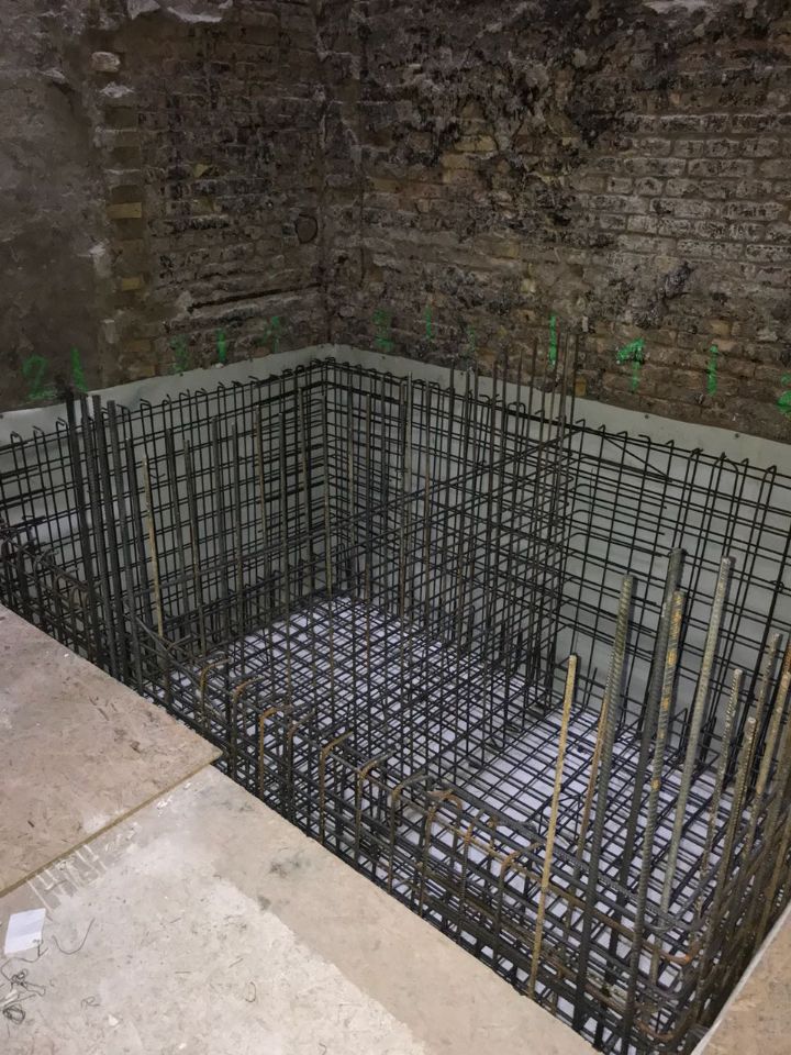 На этом изображении показан участок строительства, который кажется незаконченным. В центре снимка находится железный каркас в форме ящика, скорее всего, предназначенный для заливки бетона. Каркас сделан из железной проволоки с покрытием и имеет несколько выступающих вертикально железных прутьев. Вокруг каркаса находится пол, который, как представляется, сделан из другого материала, с участком, который слева кажется более гладким. Стены на заднем плане выглядят, возможно, неухоженным кирпичом, с некоторыми вкраплениями и неравномерным цветом. Также есть следы зеленой окраски, которые указывают на определенный размер или ограничение стены.