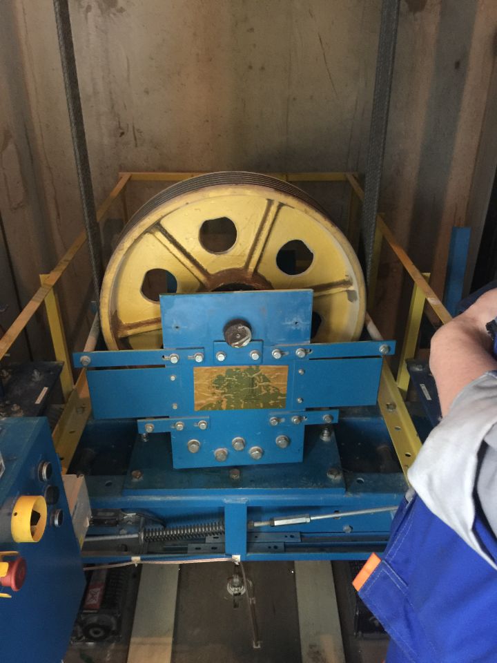 На этом изображении показана механическая система, которая, скорее всего, используется в лифте или подъемном устройстве. К тросу подключено большое желтое колесо. Эти колеса, по-видимому, служат для подъема или опускания тяжестей. В центральной части находится конструкция синего цвета, имеющая несколько болтов и шурупов, указывающих на то, что это важная часть подъемного механизма. С правой стороны вы можете видеть руку человека в синей рабочей одежде, который, возможно, контролирует или обслуживает устройство. Фон виден на фоне бетонных стен. Общая атмосфера отражает типичную рабочую среду данной техники.