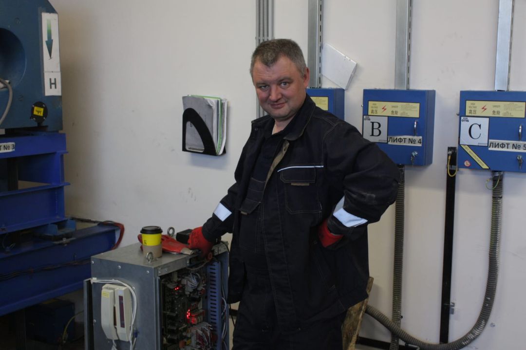 На изображении мужчина в рабочем комбинезоне и перчатках стоит, по-видимому, в промышленной или ремонтной зоне. Его комбинезон тёмного цвета, возможно, тёмно-синего или чёрного, со светоотражающими полосками на рукавах, что указывает на то, что это, скорее всего, защитная или яркая одежда, которая обычно используется в промышленных условиях. Одна его рука лежит на бедре, а другая — на открытом электрическом или механическом блоке управления, в котором видны провода и компоненты. Его поза и выражение лица говорят о том, что он уверен в себе и, возможно, немного шутит или гордится своей работой. Справа от него (слева на изображении) находится металлический корпус или механизм с синей внешней поверхностью, возможно, часть оборудования, которое он обслуживает. На стене позади него установлены синие панели управления с надписями на кириллице. На каждой панели есть большая буква, например «B» и «C», а также надписи «ЛИФТ» (по-русски «лифт» или «подъёмник») и соответствующие номера, указывающие на то, что это помещение может быть кабинетом управления лифтом или механической комнатой, связанной с работой лифтов. Жёлтые предупреждающие символы на этих коробках указывают на высокое напряжение, подчёркивая требования безопасности на рабочем месте. На стене также висит подставка для документов, в которой лежат какие-то руководства или журналы, часто используемые для записей о техническом обслуживании. Кроме того, на открытой панели управления стоит чашка с кофе, что придаёт обстановке индивидуальность и намекает на перерыв или неформальную рабочую обстановку. В целом, это изображение реалистично передаёт образ промышленного рабочего в технической среде, демонстрируя элементы работ по техническому обслуживанию в лифте или электрощитовой.