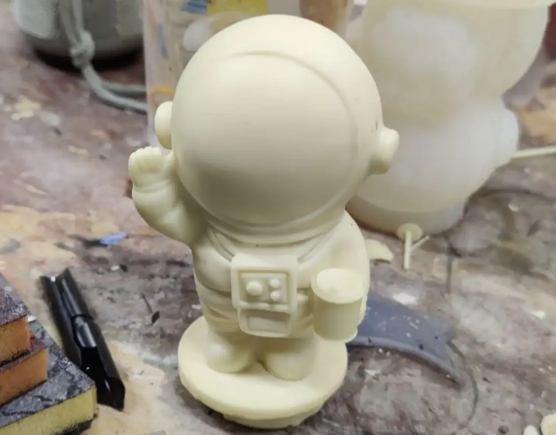 Литая фигурка космонавта без окраски, стоящая на рабочем столе рядом с инструментами и формой.