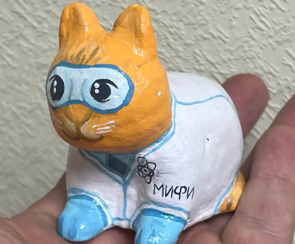 Ручная фигурка кота-учёного с очками и халатом, сфотографированная на фоне стены.
